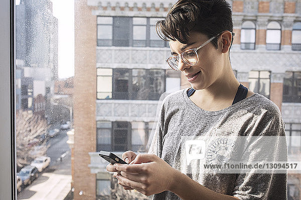 Frau mit kurzen Haaren benutzt Mobiltelefon  während sie am Fenster steht
