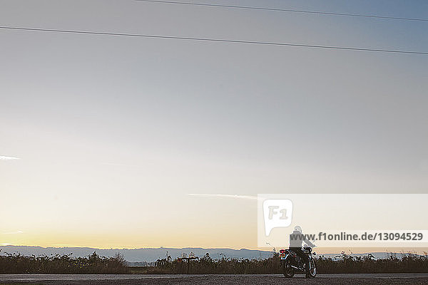 Rear view of male biker sitting on bike at road against sky during sunset