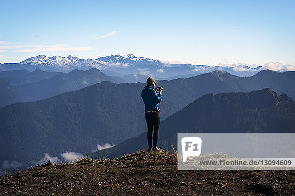 Rückansicht einer Frau  die an einer Klippe vor Bergen fotografiert