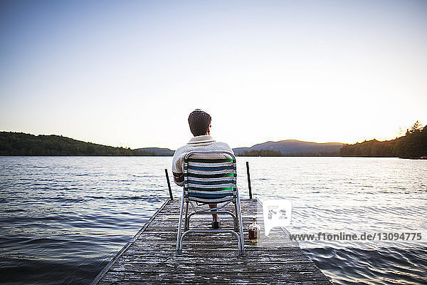 Rear view of man sitting on deck chair on jetty by lake