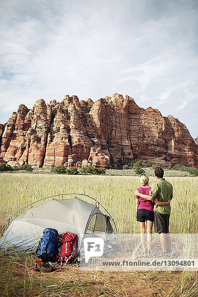 Rear view of couple standing by tent at grassy field against mountain