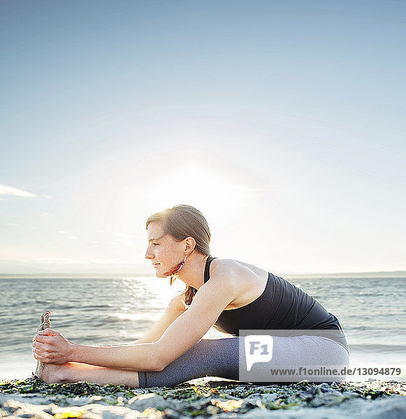 Woman practicing seated forward bend at beach against sky