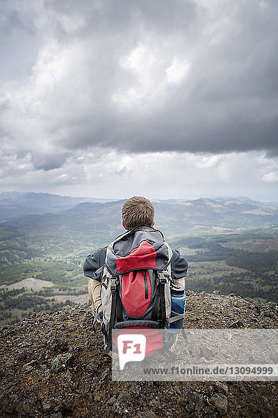 Rückansicht eines Wanderers mit Rucksack  der auf einem Berg vor bewölktem Himmel sitzt