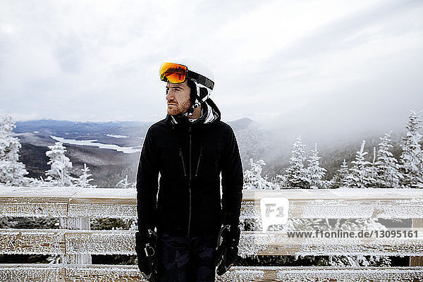 Nachdenklicher Mann in Skikleidung  der bei nebligem Wetter gegen einen gefrorenen Holzzaun steht