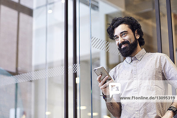 Glücklicher Geschäftsmann schaut weg  während er ein Smartphone im Büro hält