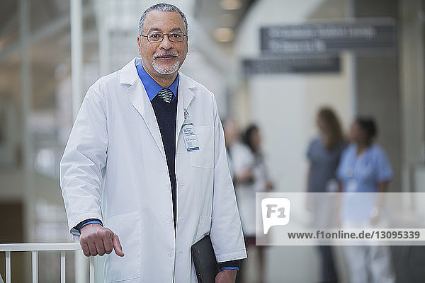 Porträt eines männlichen Arztes  der im Korridor steht  während weibliche Mitarbeiter im Hintergrund diskutieren