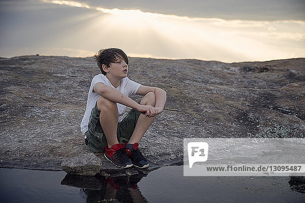 Junge schaut weg  während er am Bach sitzend auf dem Arabien-Berg gegen bewölkten Himmel