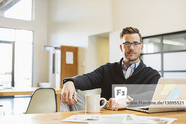 Portrait of businessman with laptop computer and coffee cup sitting at desk in office