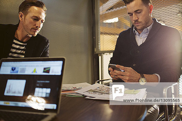 Geschäftsmann benutzt Mobiltelefon  während er mit einem männlichen Kollegen im Büro sitzt