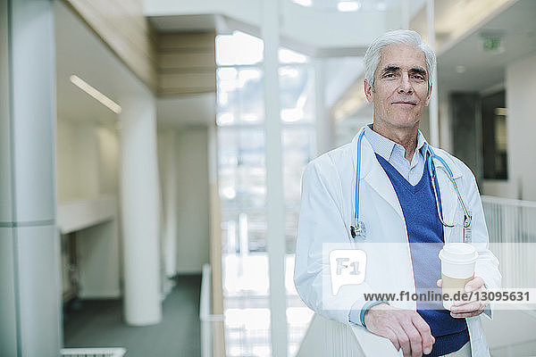Portrait of male doctor holding disposable cup while standing in hospital corridor