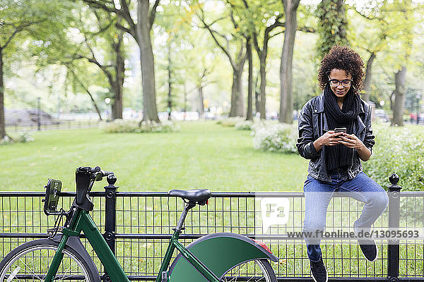 Frau mit Fahrrad benutzt Mobiltelefon  während sie im Park auf einem Geländer sitzt