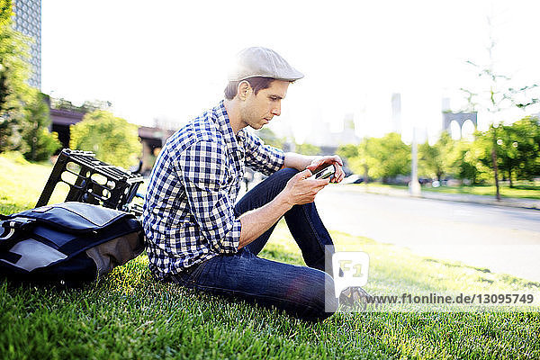 Man using smart phone while sitting on grass field against clear sky