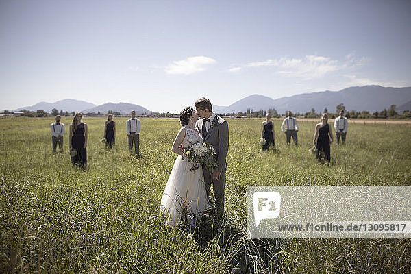 Neuvermähltes Paar küsst sich  während es bei sonnigem Wetter auf einem Grasfeld steht  mit Trauzeugen und Brautjungfern im Hintergrund