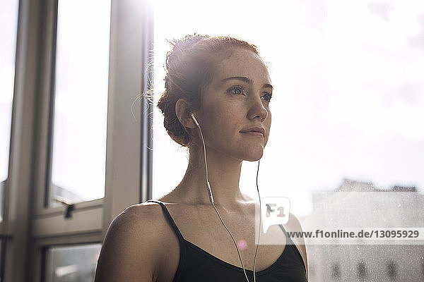 Low angle view of thoughtful woman listening music while standing by window at home