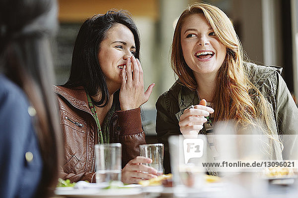 Glückliche Frau flüstert am Tisch im Restaurant in das Ohr eines Freundes