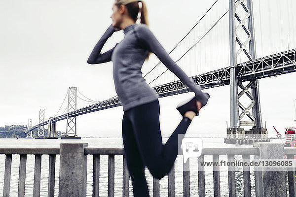 Seitenansicht einer Athletin  die auf einem Fußweg ihr Bein streckt  mit der Oakland Bay Bridge im Hintergrund