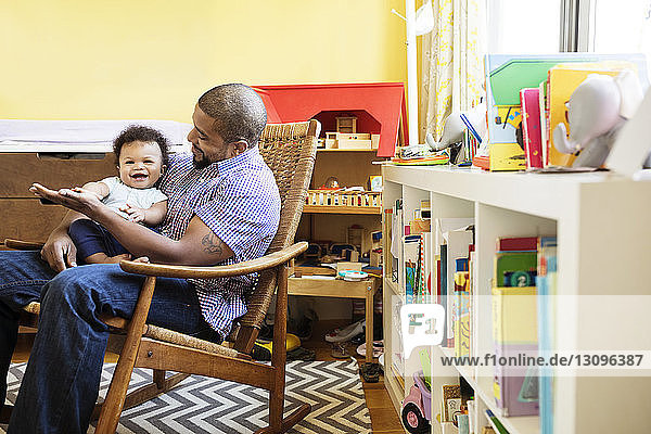 Vater spielt mit dem Sohn  während er zu Hause auf einem Sessel sitzt