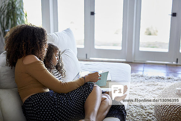 Hochwinkelansicht einer Mutter  die ein Bilderbuch für ihre Tochter liest  während sie zu Hause auf dem Sofa sitzt