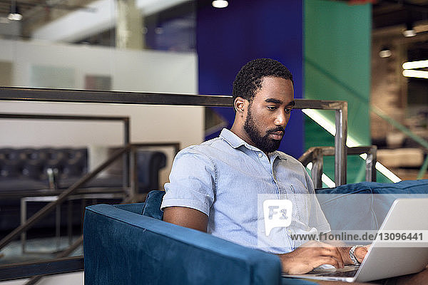 Seriöser Unternehmer benutzt Laptop  während er im Kreativbüro auf dem Sofa sitzt