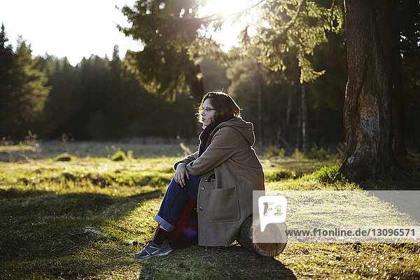 Seitenansicht einer nachdenklichen Frau  die wegschaut  während sie auf einem Baumstamm im Wald sitzt