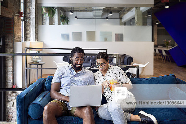 Glückliche Geschäftsleute schauen auf den Laptop  während sie im Kreativbüro auf dem Sofa sitzen