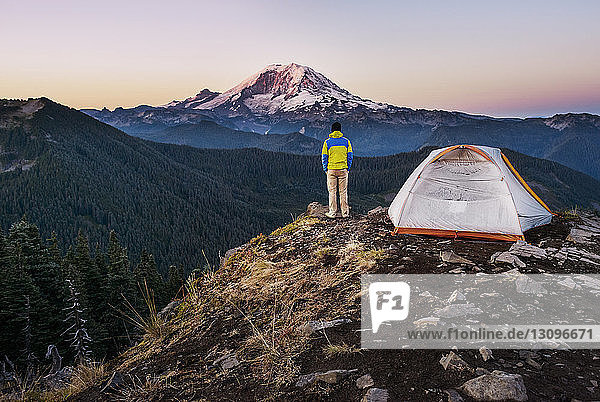 Rückansicht eines Mannes  der am Zelt auf einer Klippe vor den Bergen steht