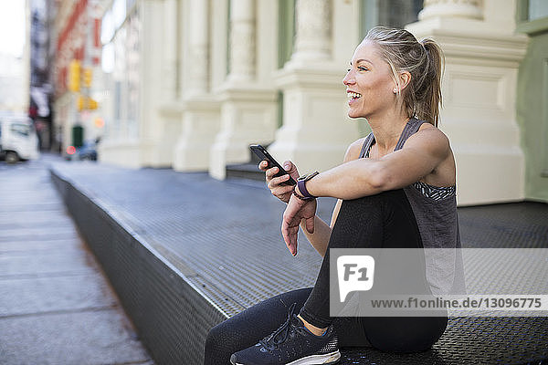 Glücklicher Sportler benutzt Mobiltelefon  während er in der Stadt auf dem Bürgersteig sitzt