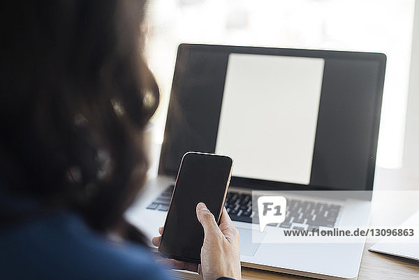 Ausgeschnittenes Bild einer Geschäftsfrau  die ein Mobiltelefon benutzt  während sie im Büro an einem Laptop-Computer sitzt