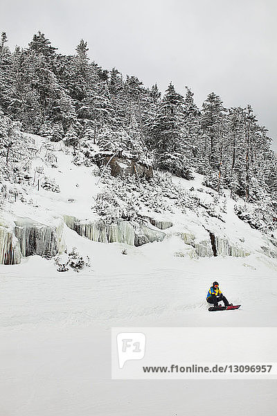 Mann sitzt mit Snowboard auf schneebedecktem Feld