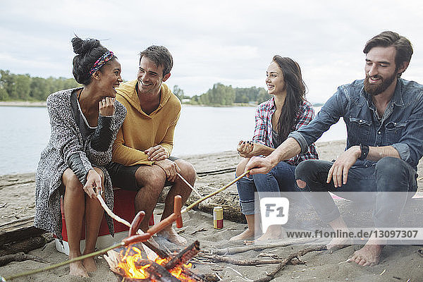 Happy friends roasting sausages while sitting at campfire against river