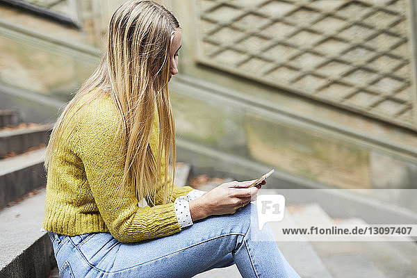 Seitenansicht einer jungen Frau  die ein Mobiltelefon benutzt  während sie auf einer Treppe sitzt