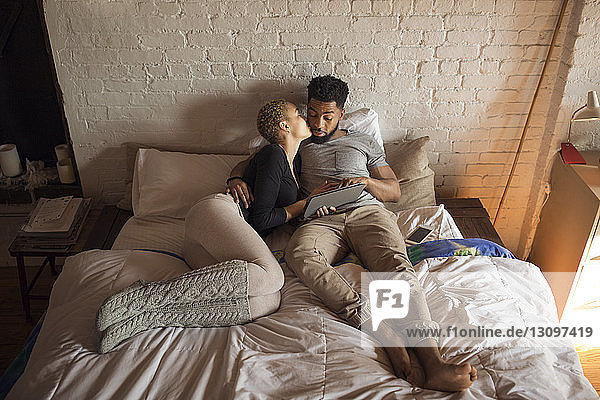 Draufsicht einer romantischen Frau  die einen Mann küsst  während sie einen Tablet-Computer am Bett benutzt