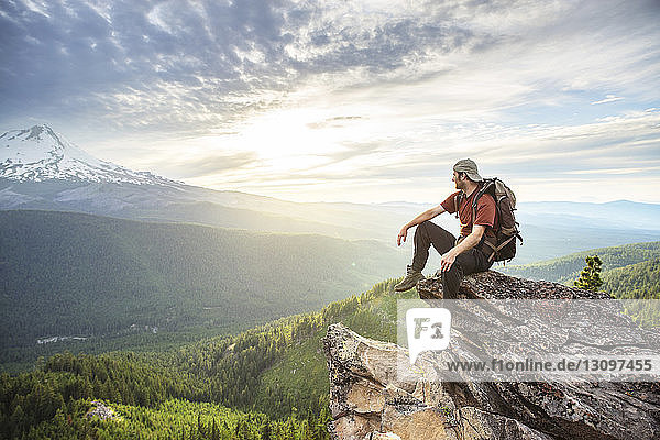 Hiker looking away while sitting on mountain against cloudy sky