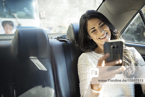 Glückliche junge Frau  die sich im Taxi durch ein Smartphone selbstständig macht
