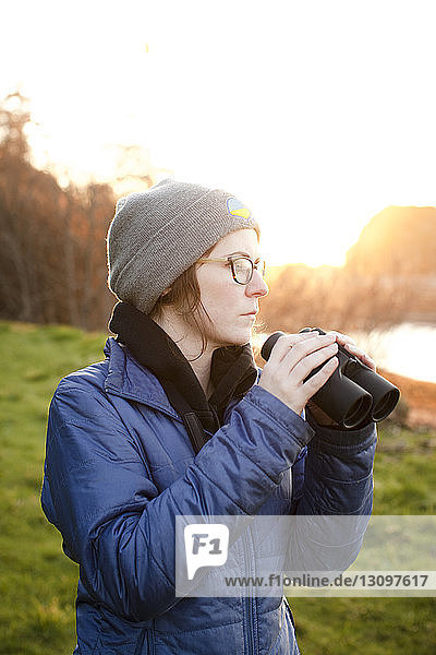 Woman holding binoculars looking away while standing on grassy field
