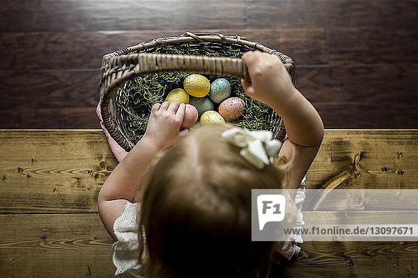 Draufsicht eines Mädchens mit einem Korb mit Ostereiern  das zu Hause auf einem Holztisch sitzt