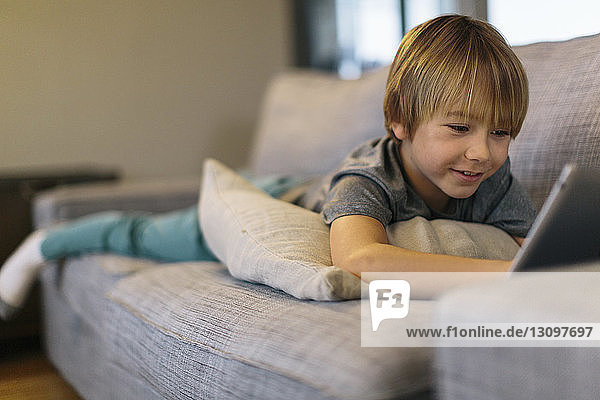 Lächelnder Junge benutzt Tablet-Computer  während er zu Hause auf dem Sofa liegt