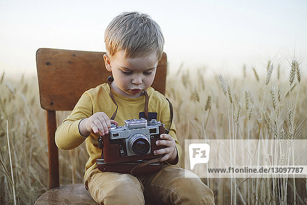 Cute boy with vintage camera sitting on chair amidst wheat field