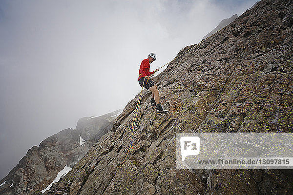 Side view of hiker using rope while climbing rock formations against clouds