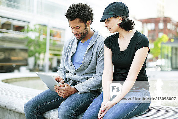 Multi-ethnic couple using tablet computer while sitting on retaining wall