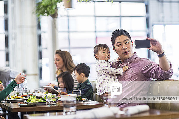 Father taking selfie with baby girl while sitting with family in restaurant