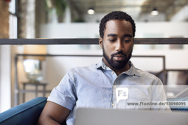 Junger selbstbewusster Geschäftsmann mit Laptop  während er im Kreativbüro auf dem Sofa sitzt