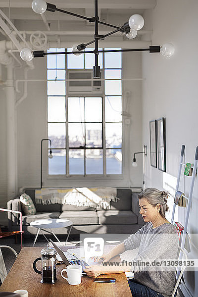 Konzentrierte reife Frau mit Laptop bei der Arbeit am Tisch im Wohnzimmer