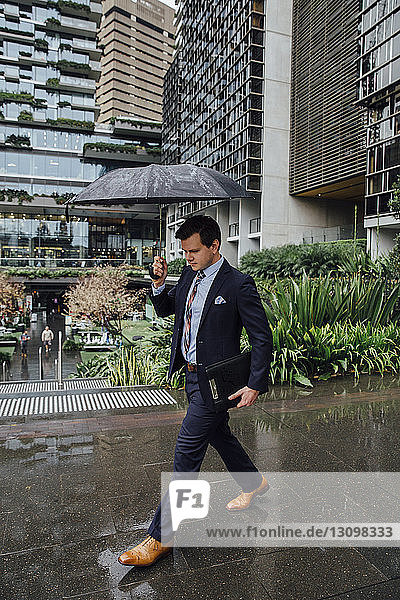 Geschäftsmann geht mit Regenschirm in der Hand auf nassem Fußweg in der Stadt