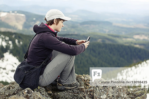 Seitenansicht einer Frau  die ein Mobiltelefon benutzt  während sie auf dem Gipfel eines Berges sitzt