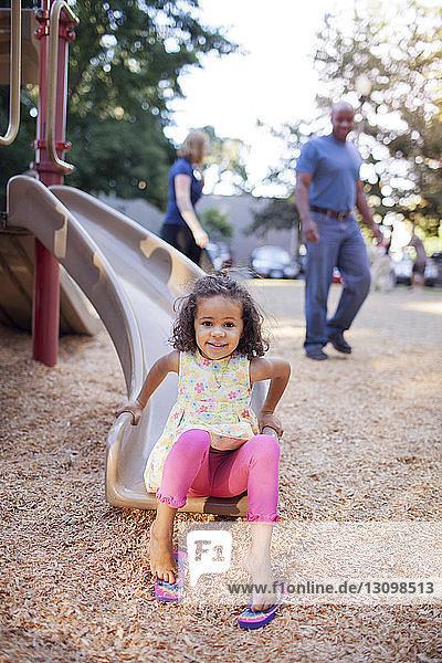 Portrait of girl sitting on slide with parents in background