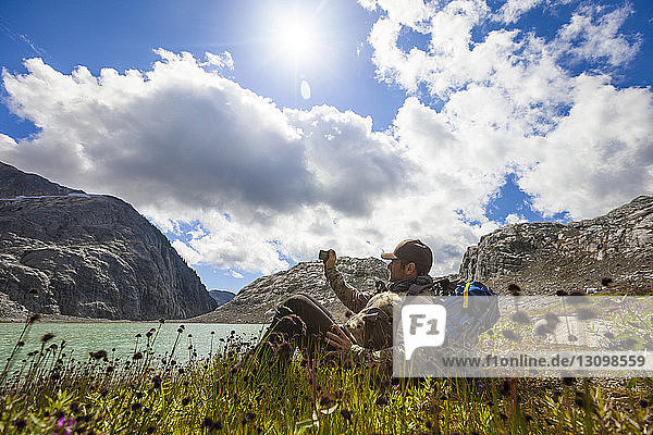 Side view of hiker photographing while relaxing on lakeshore against sky