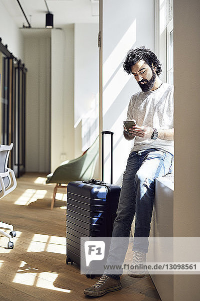 Geschäftsmann in voller Länge  der ein Smartphone benutzt  während er im Kreativbüro auf dem Fensterbrett sitzt