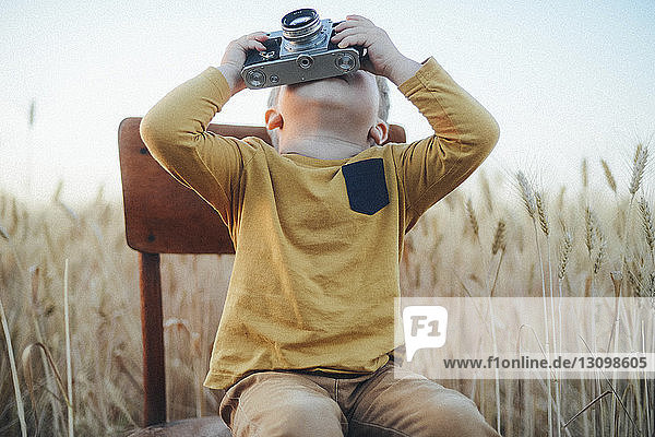 Playful boy photographing with vintage camera while sitting on chair amidst wheat field