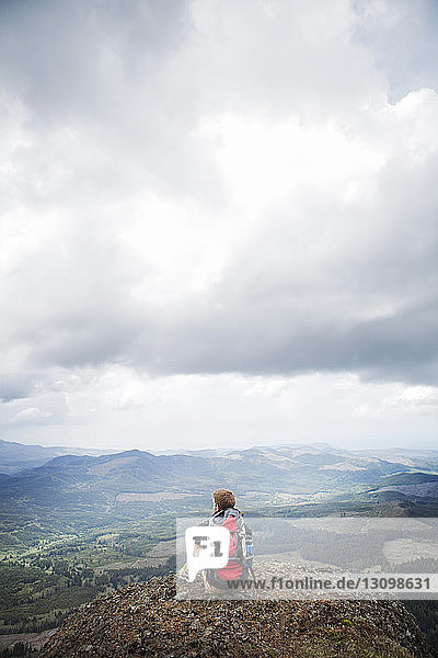 Hiker with backpack sitting on mountain against cloudy sky